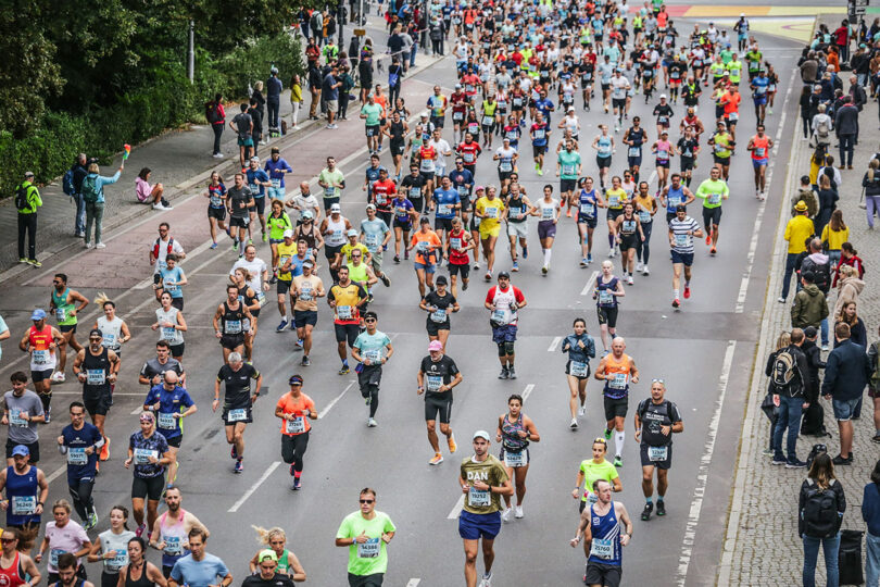 Amanda Gunawan joins a large group of runners participating in a city marathon on a wide road, surrounded by enthusiastic spectators and lush greenery.