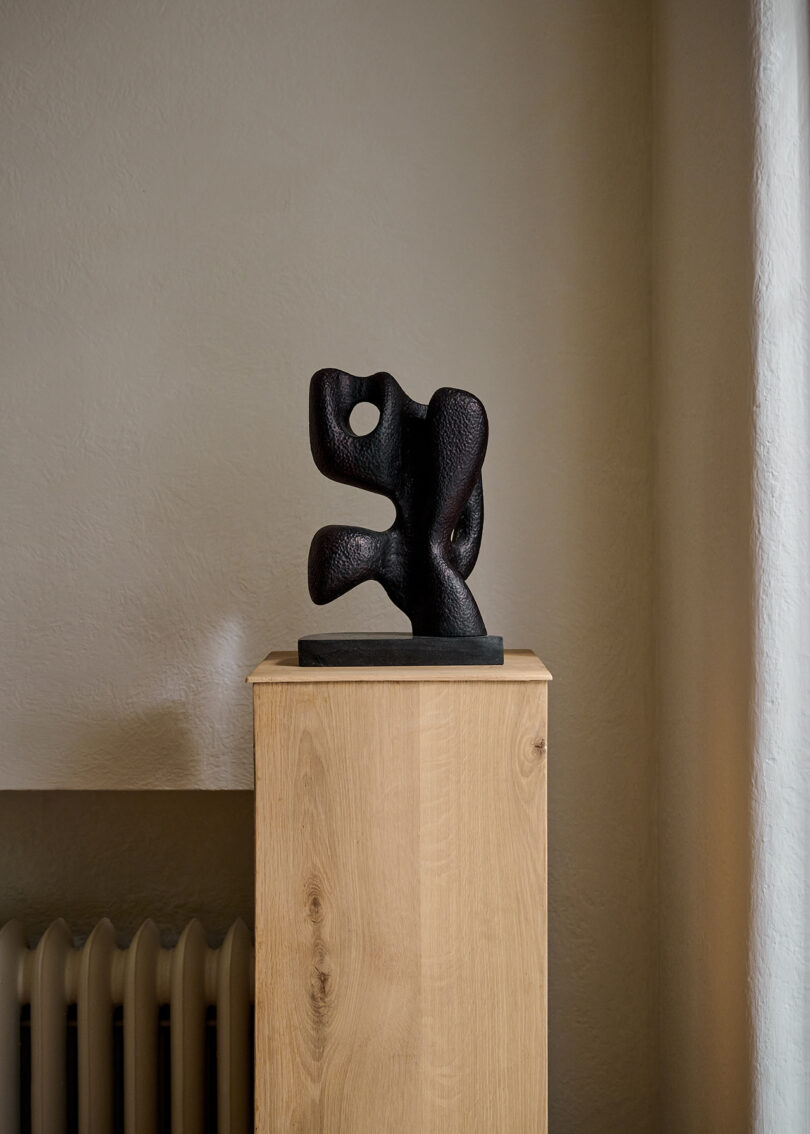 Abstract black sculpture on a wooden pedestal against a plain wall, with a radiator partially visible on the left.