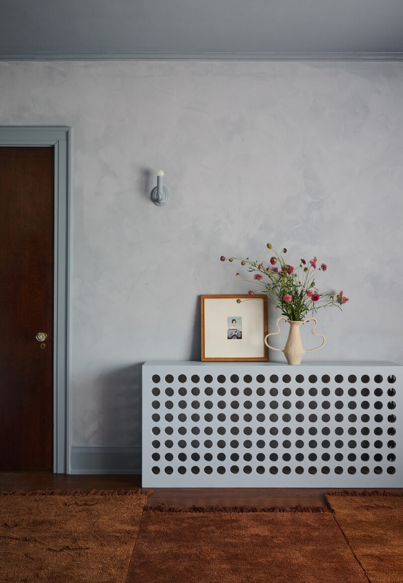 A decorated console with a perforated front holds a framed picture and a vase of flowers against a textured, pale blue wall. A wooden door and a sconce light are visible.