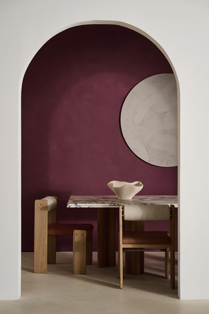 Modern dining area with a wooden table, two chairs, a white ceramic bowl, and a circular textured wall art against a maroon accent wall, framed by a white archway.