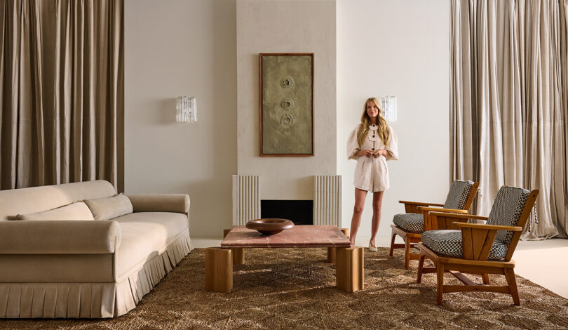 A woman stands in a living room with a beige sofa, two wooden chairs, a red stone coffee table, and a painting on a cream wall. Heavy drapes frame two tall windows.