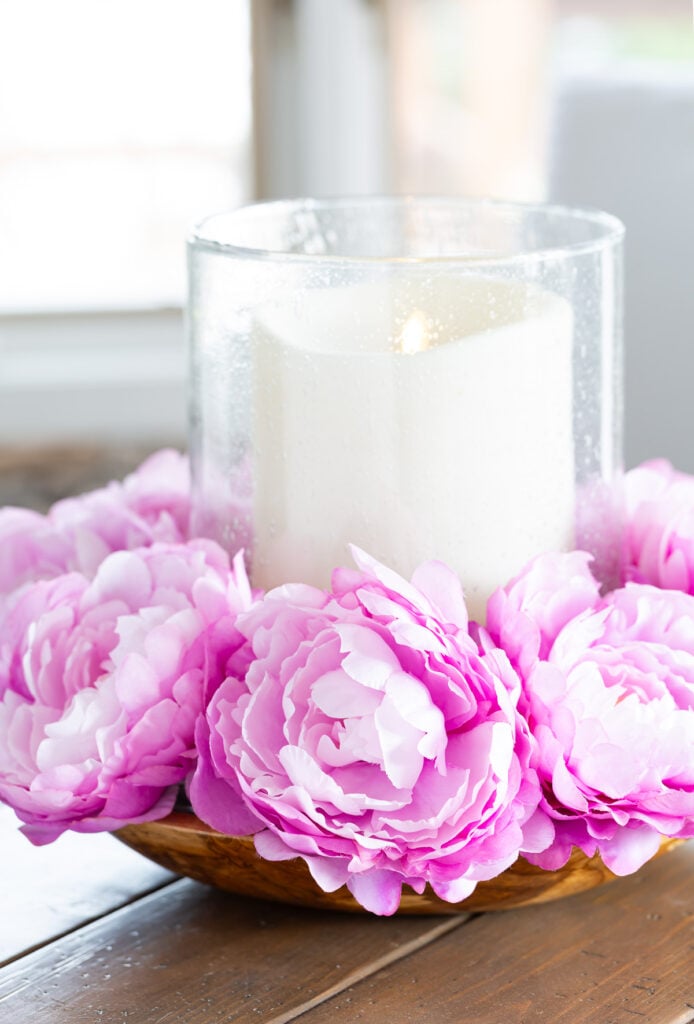 Pink flowers surrounding a large candle that is placed in a large bowl on a wood table.