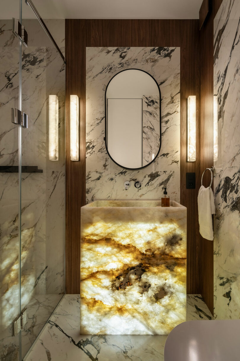 Luxurious bathroom with marble walls, a lighted onyx sink, oval mirror, and vertical light fixtures. Glass shower on the left with a dark wood accent wall framing the wash area.