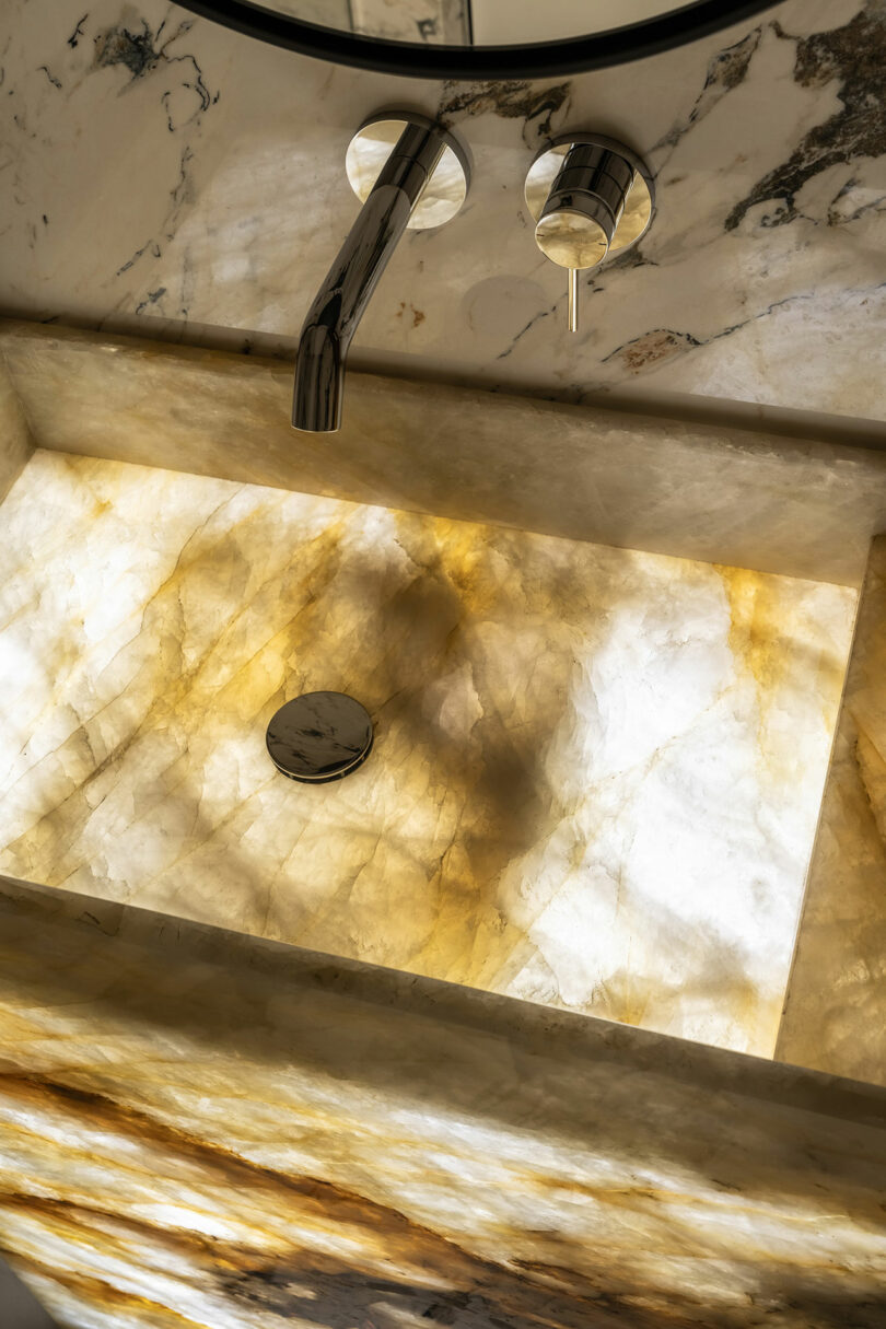 Illuminated marble sink with a chrome faucet and soap dispenser, set against a marbled wall.