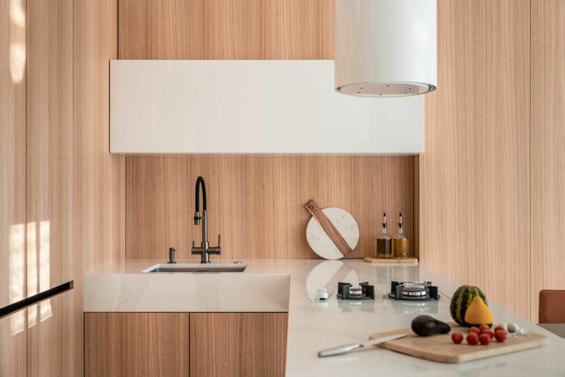 Modern kitchen with wooden cabinets, white countertop, and appliances. A cutting board with vegetables and a stovetop are on the island. Brown wall panels with integrated lighting are shown.