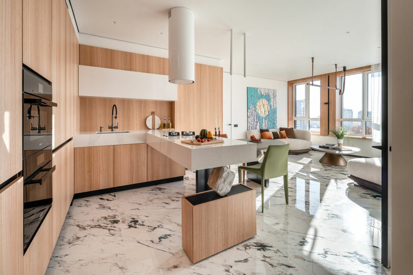 Modern kitchen and living area with light wood cabinets, marble floor, and large windows. Features a central island, dining chairs, and artwork on the wall.