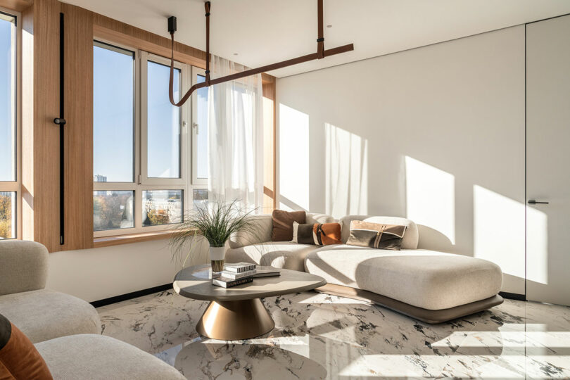 Modern living room with large windows, light walls, a plush sectional sofa, and a round marble coffee table. Sunlight streams in, highlighting the minimalist decor and wooden accents.