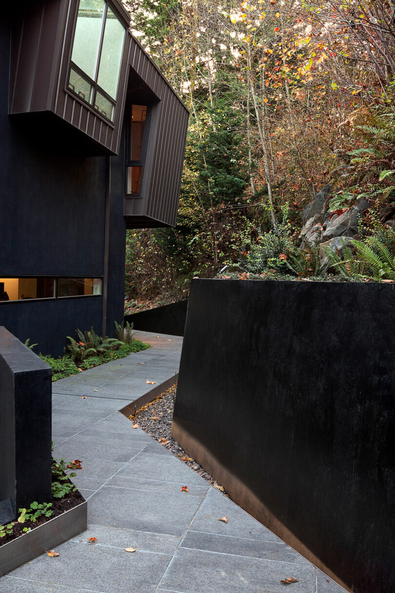 Modern house exterior with dark walls and large windows, surrounded by lush greenery and a narrow stone pathway leading alongside the building.