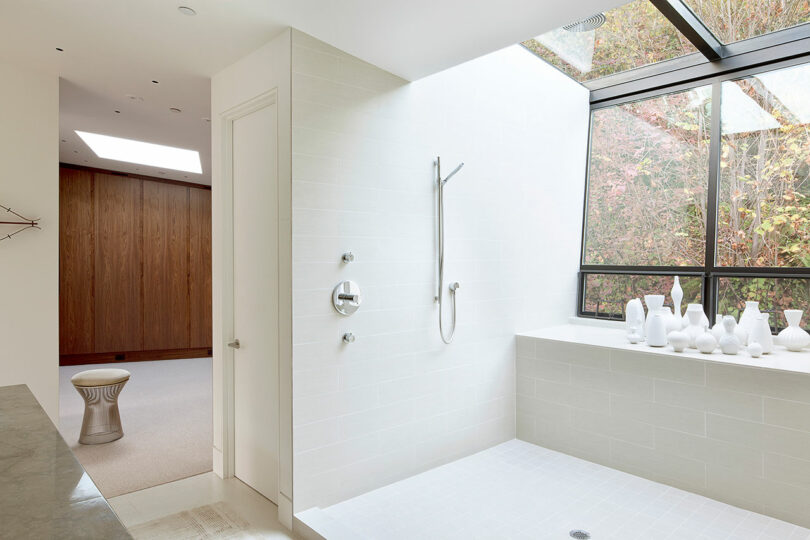 A bright bathroom with a walk-in shower and a sloped glass roof. White tiles cover the shower walls. Various white decorative vases are displayed on the windowsill.