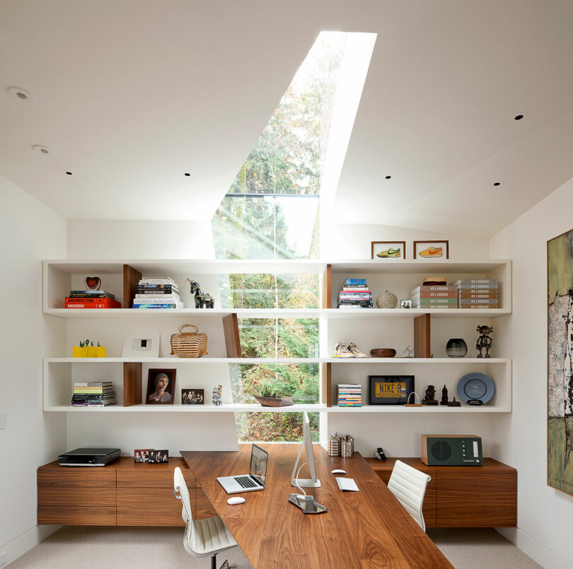 Modern home office with a wooden desk, computer, and chair. Wall-mounted shelves display books and decorative items. A skylight above illuminates the space, and a window shows greenery outside.