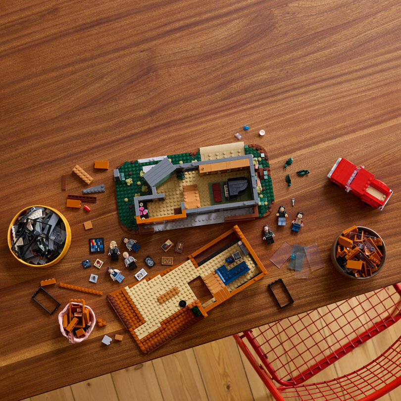 A wooden table with LEGO pieces, several minifigures, and partially assembled structures. A red wire chair is partially visible in the corner.