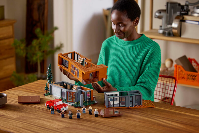 Person in green sweater assembling a LEGO house set with various pieces and minifigures on a table.