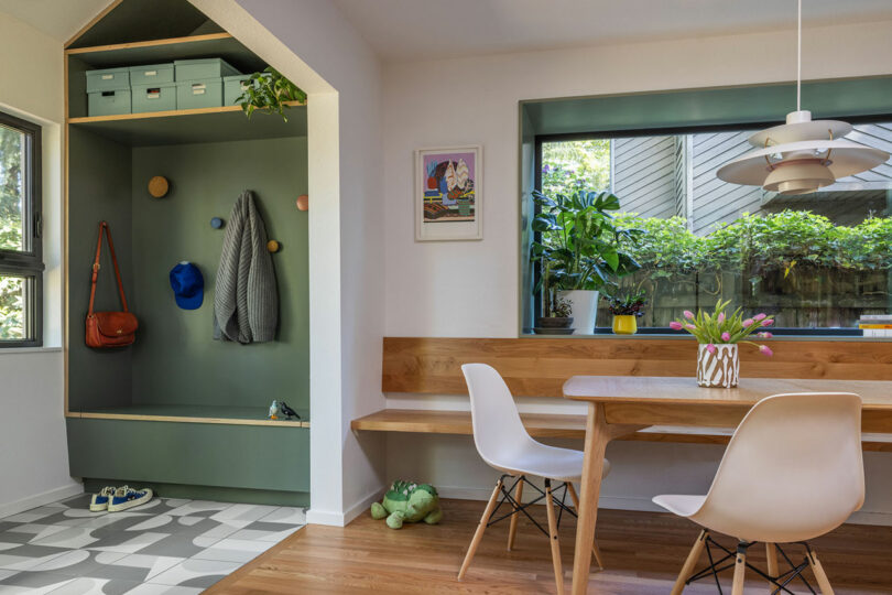 A cozy dining area with a wooden table, white chairs, and a window seat with plants. A built-in green cubby with hooks, shelves, and a bench is nearby. Natural light brightens the space.