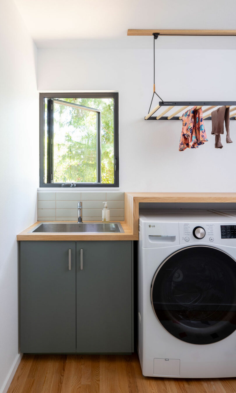 A laundry room with a sink, countertop, and a front-loading washing machine. A wooden drying rack above holds clothes. An open window allows natural light inside.