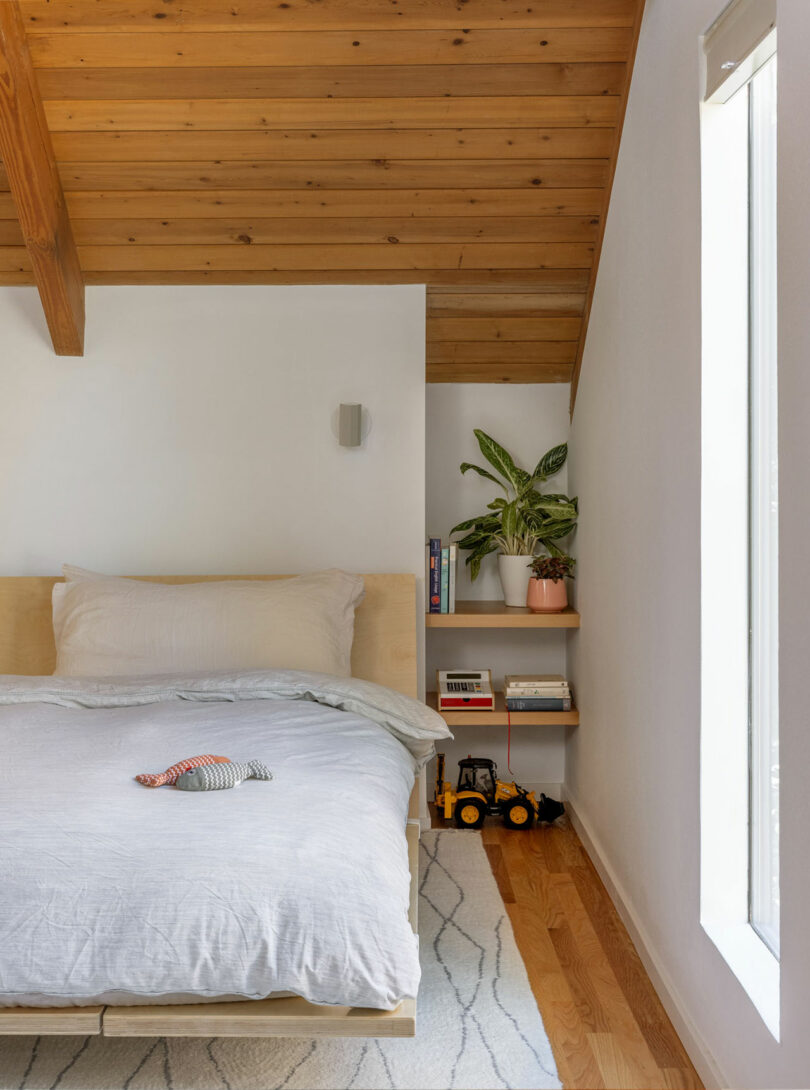 Cozy bedroom with wooden ceiling, bed with light gray bedding, stuffed toy, side shelves with plant and books, small construction toys on floor, and a window.