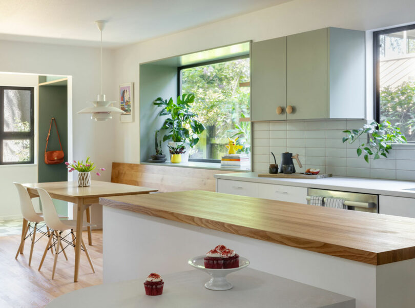 Modern kitchen with light green cabinets, wooden countertops, large windows, and indoor plants. A table with chairs and a counter with two cupcakes are in the foreground.