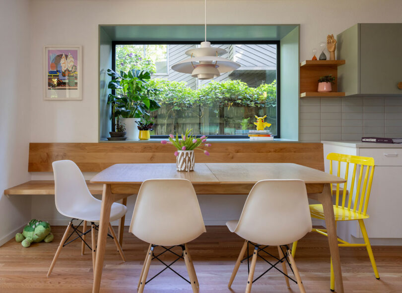 Modern dining room with a wooden table, white and yellow chairs, a window view of greenery, and plants on the sill. Pendant light hangs above.