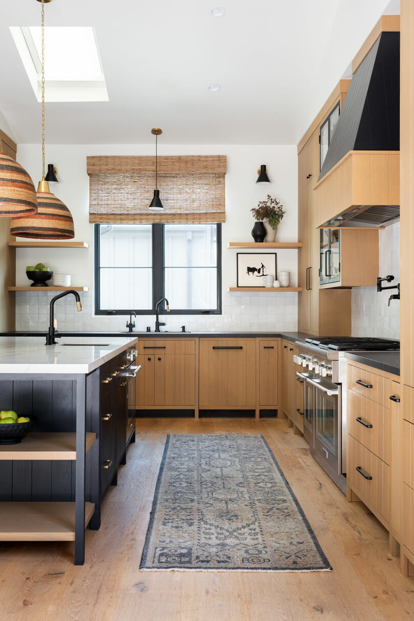 A modern kitchen with wooden cabinets, a central island, a rug, and two hanging lights. A window with a woven blind and various kitchen appliances are visible.