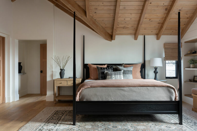 A bedroom with a wooden canopy bed, gray and orange bedding, a wooden nightstand with a vase, a lamp, and a woven rug on the wooden floor beneath a slanted wooden ceiling.