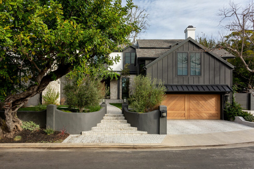 Modern two-story house with dark gray exterior, wooden garage door, and stone pathway. Surrounded by trees and shrubs, the entrance features wide steps leading to a wooden main door.