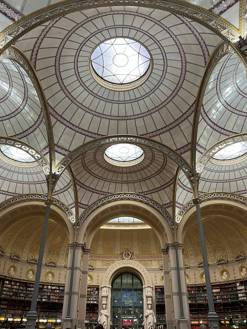 Circular domed ceiling with intricate metalwork and glass, reminiscent of Elizabeth Graziolo's refined designs, crowns an ornate library interior. Arched doorways and tall shelves of books stand below, creating a timeless sanctuary for knowledge.