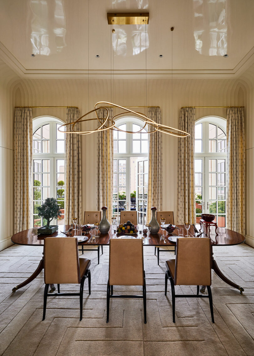 Elegant dining room designed by Elizabeth Graziolo, featuring a large wooden table, eight chairs, decorative vases, a curved light fixture, and tall windows adorned with patterned curtains.