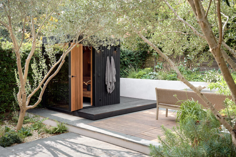 Small wooden sauna with a towel hanging on the door, surrounded by trees and greenery, next to a wicker bench on a wooden deck