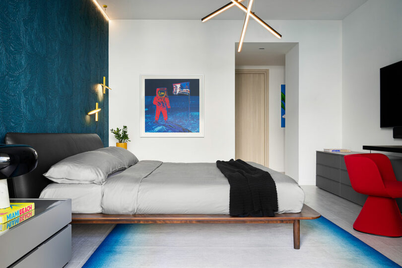 Modern bedroom with a low-profile bed, dark headboard, textured blue accent wall, astronaut artwork, and contemporary lighting. Red chair and flat-screen TV complete the minimalist decor.