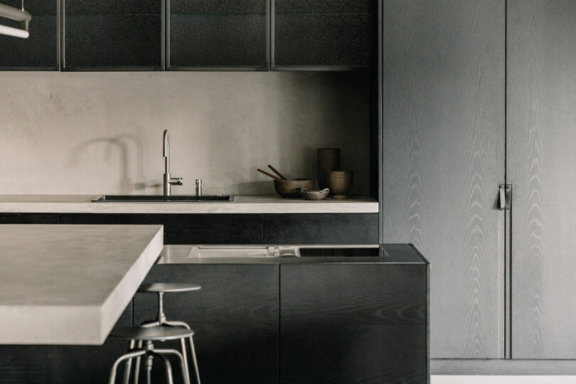 Modern kitchen with dark cabinets, light countertops, a stainless steel sink, and a metal bar stool. Minimalist decor with bowls and utensils on the counter.