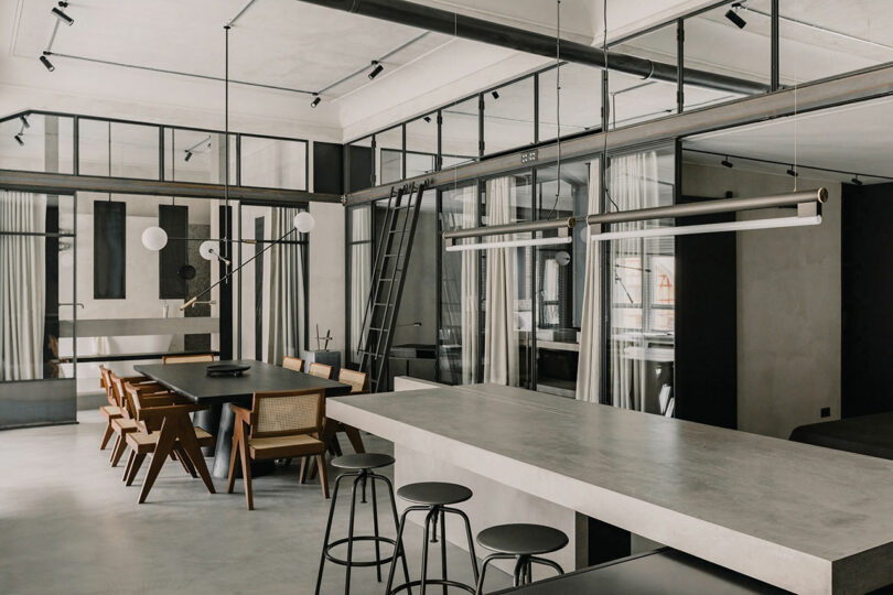 Modern industrial-style kitchen and dining area with concrete surfaces, black and wood furniture, pendant lighting, and large windows.