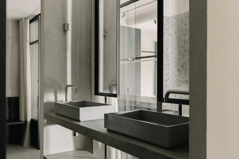 Minimalist bathroom with two rectangular sinks, large mirrors, and modern fixtures on a gray countertop. Neutral tones dominate the decor.