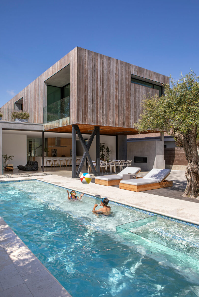 Modern two-story house with wood paneling, glass windows, and an outdoor pool. Two people swim in the pool, while a beach ball floats nearby. A patio area with seating is visible under a canopy.