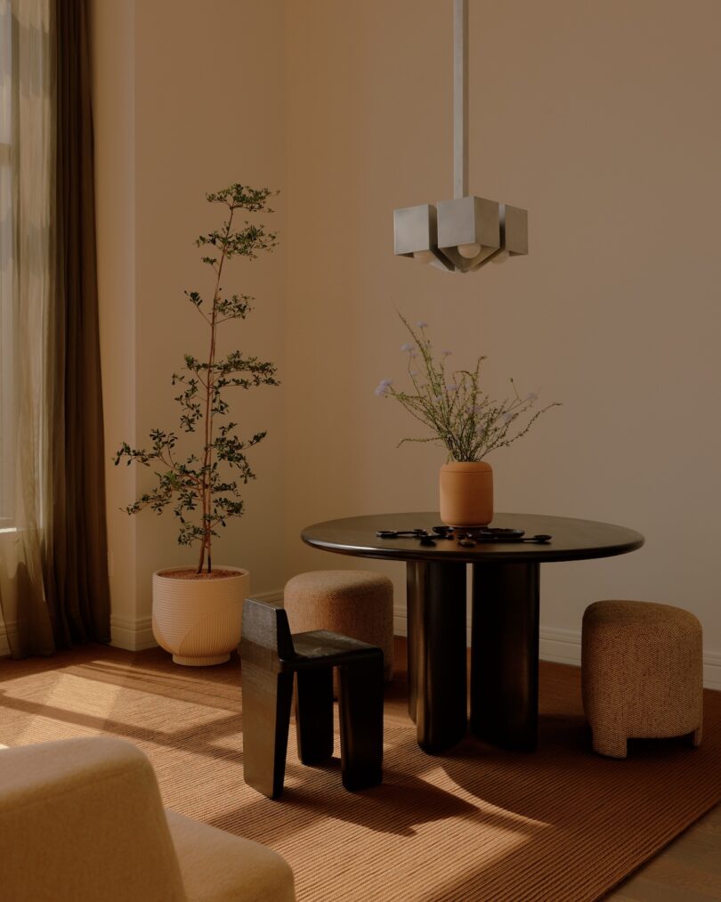 A modern dining area with a round black table, three stools, a black chair, two potted plants, a rug, and a geometric pendant light in a sunlit beige room.