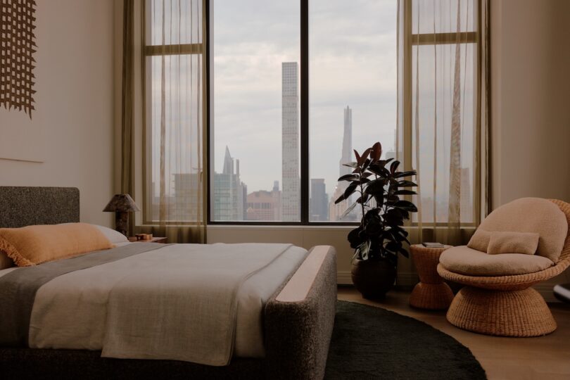 A modern bedroom with a large bed, a round woven chair, a tall plant, and floor-to-ceiling windows overlooking a city skyline.