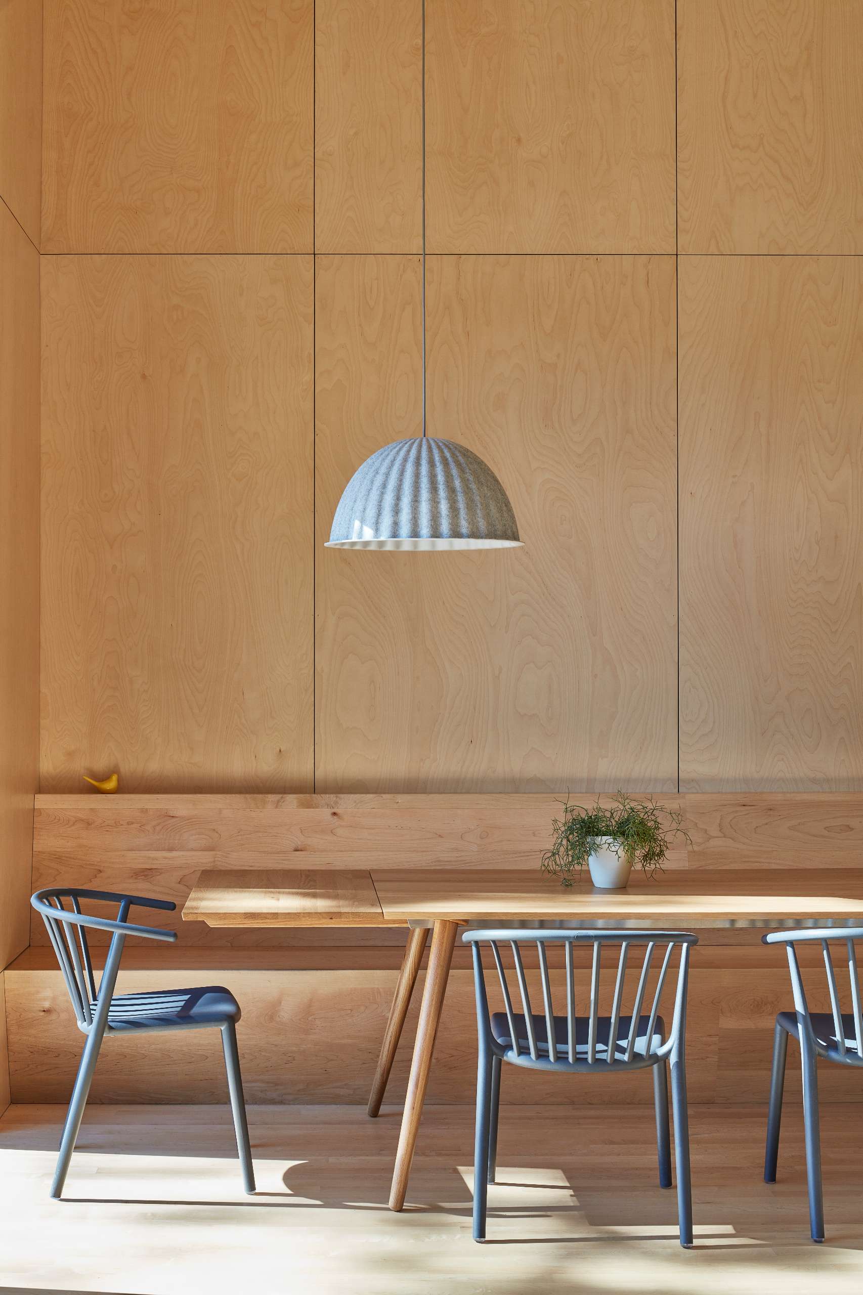 At the center of the living space in this modern cottage, the dining area includes a built-in maple plywood banquette, creating a warm and inviting atmosphere for gatherings around the dining table.