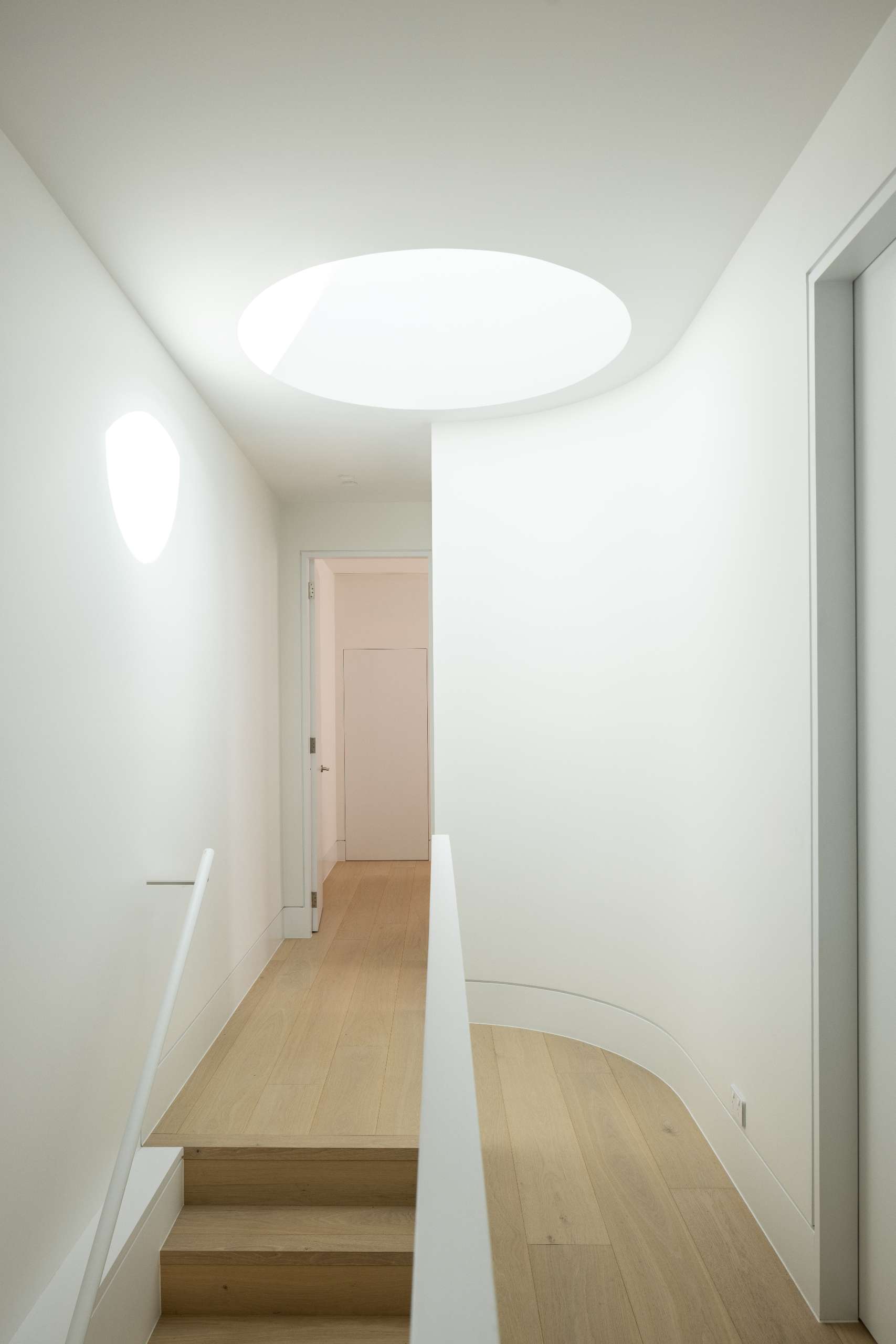 A circular skylight above the stairwell draws natural light into the heart of the home, creating a quiet moment of drama. This oculus not only improves passive solar performance but also casts shifting shadows that soften the minimalist palette and highlight the home's curved interior lines.