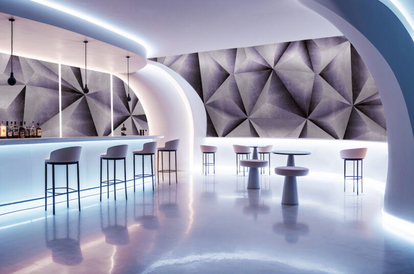 A modern bar with geometric wall art, curved white structures, sleek bar stools, and glowing blue and white lighting
