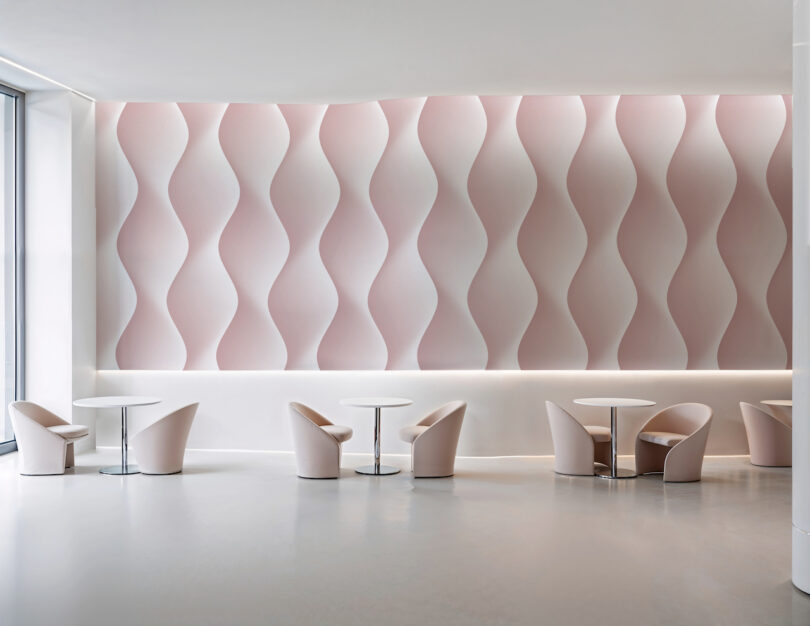 Minimalist seating area with small round tables and pale pink chairs, set against a wall with a pink and white wavy pattern. Natural light enters through large windows on the left