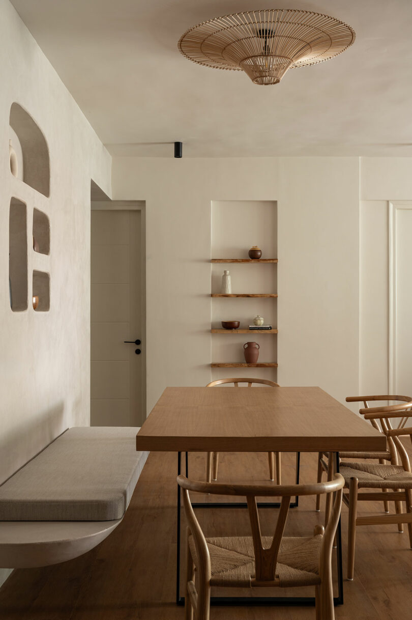 Minimalist dining room with a wooden table, four wooden chairs, a built-in bench, wall shelves with ceramics, and a woven ceiling light fixture. Neutral color palette.