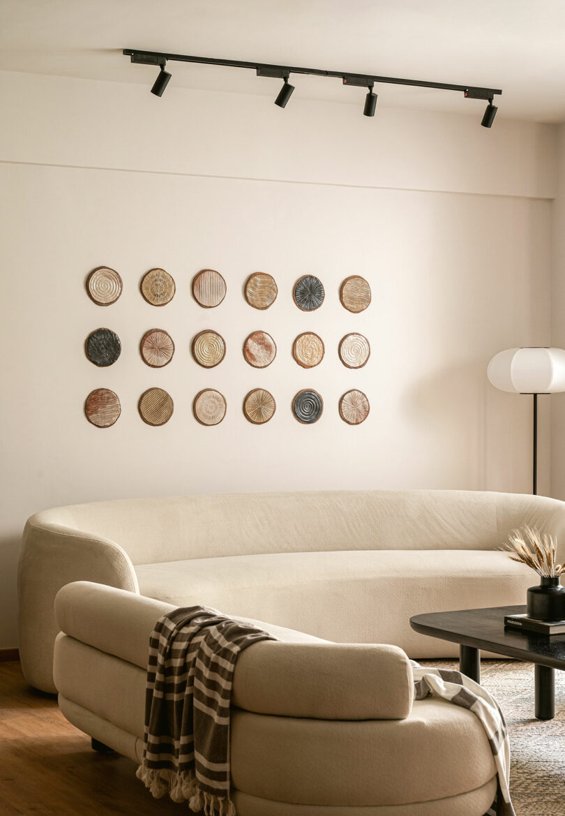 A modern living room with a curved beige sofa, a black coffee table, a floor lamp, and decorative woven plates arranged on a white wall.