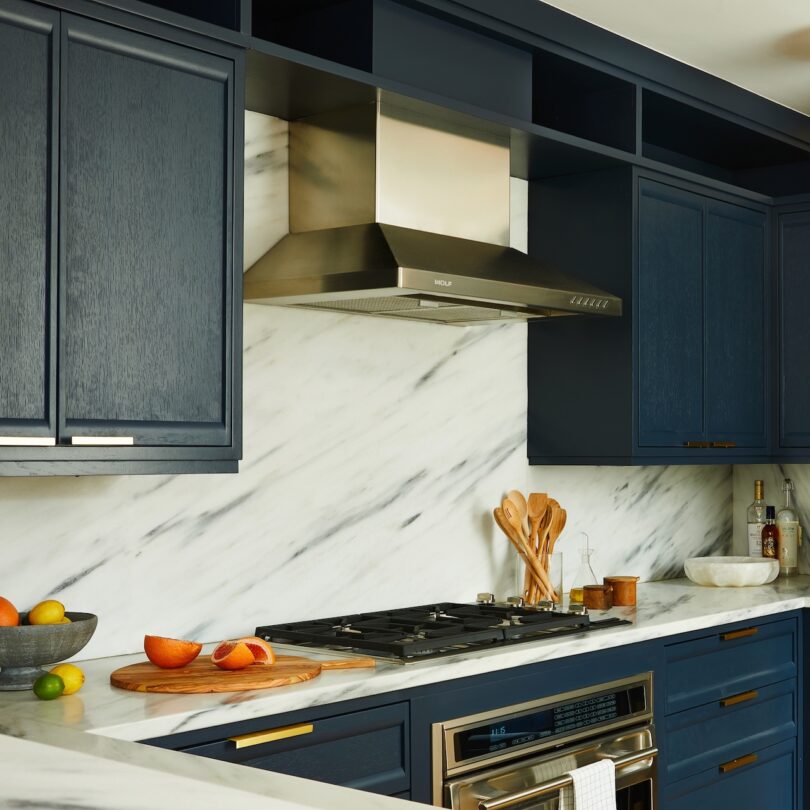 Modern kitchen with navy cabinets, marble backsplash, stainless steel range hood, and a countertop holding a cutting board with sliced carrots, utensils, and bottles.