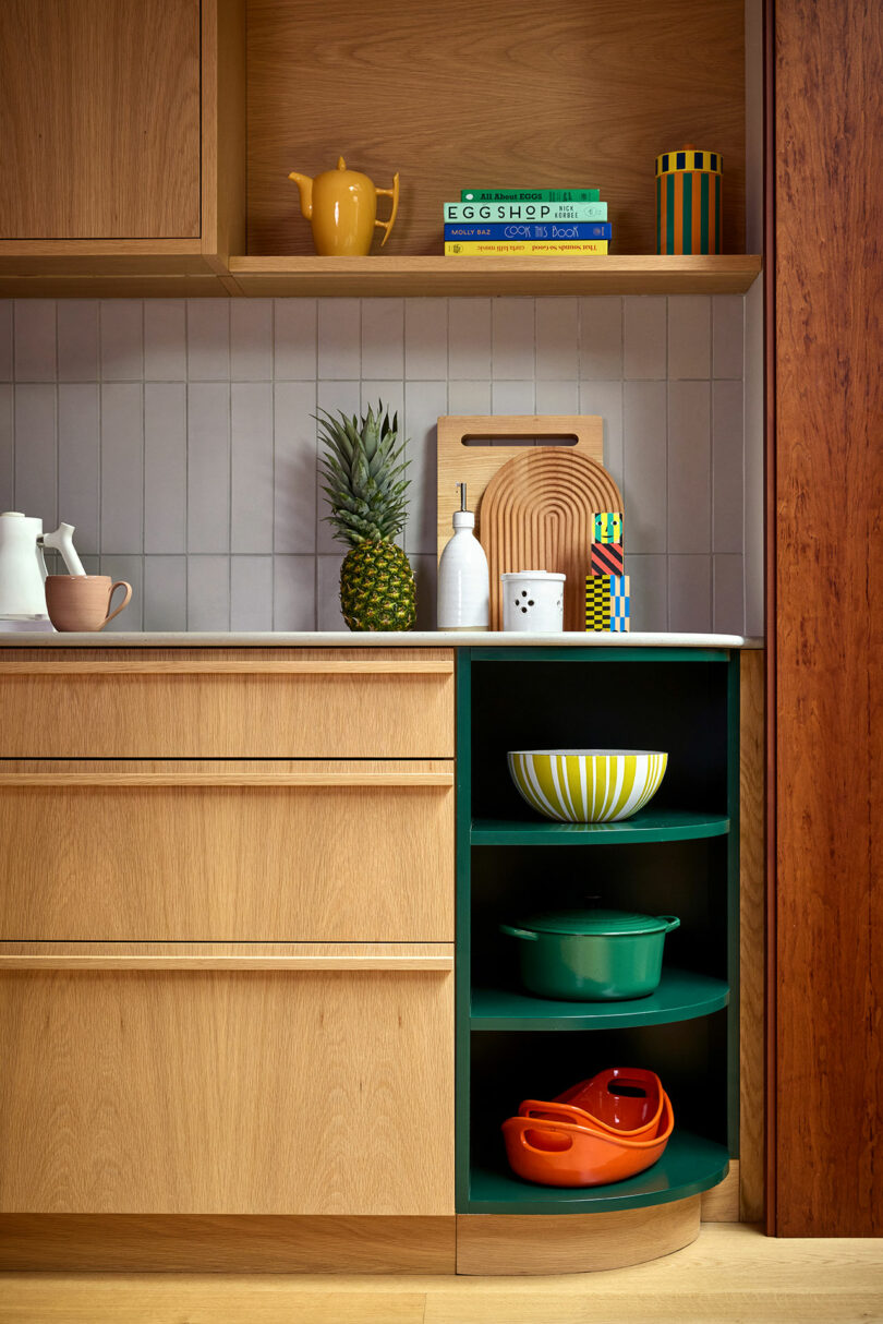 A modern kitchen corner with wooden cabinets, a pineapple, cutting boards, colorful bowls on shelves, and kitchenware arranged neatly on the counter and shelving.