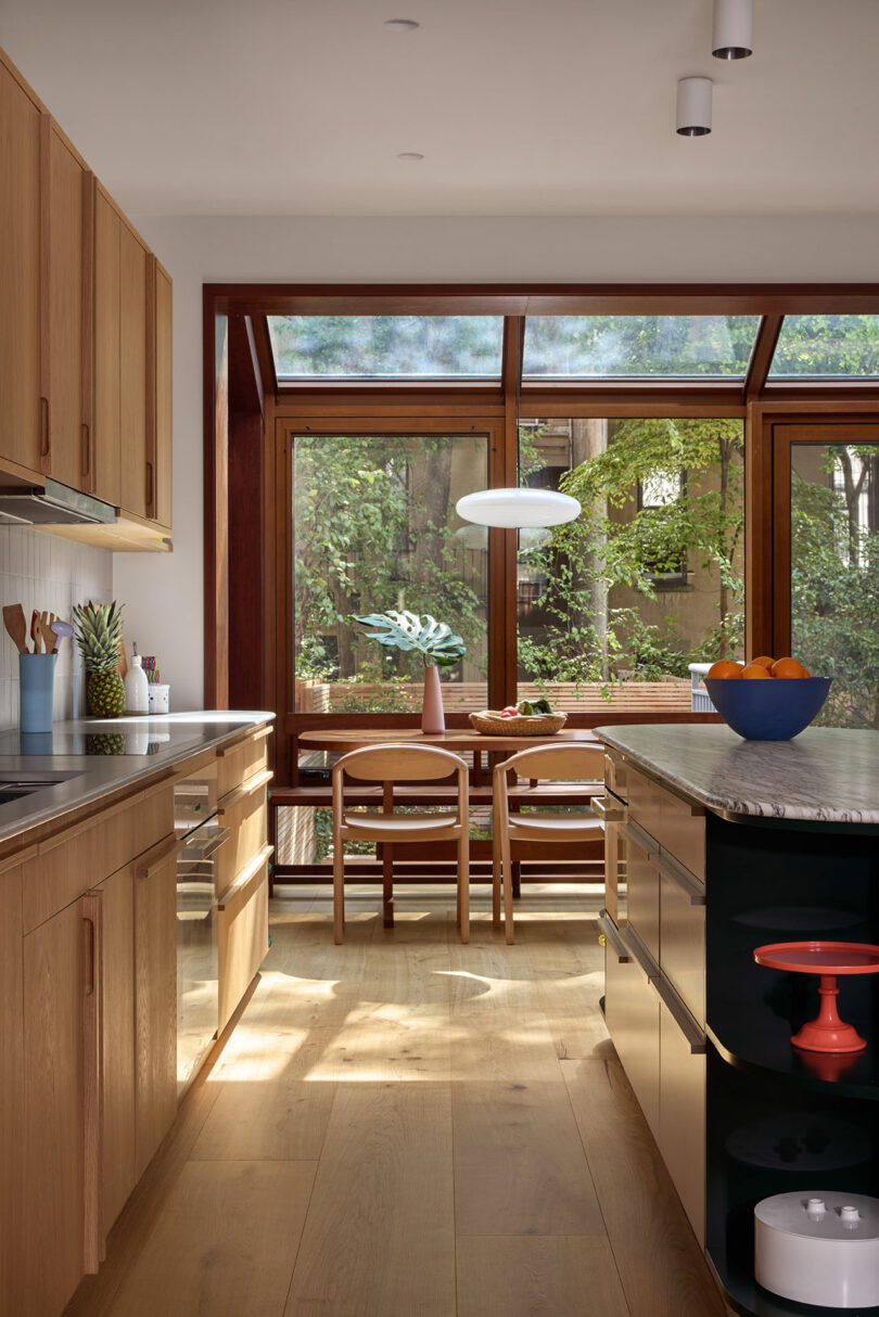 Modern kitchen with wooden cabinets, an island, and a dining area by large windows overlooking a green garden. A vase with flowers and a bowl of oranges are on display.