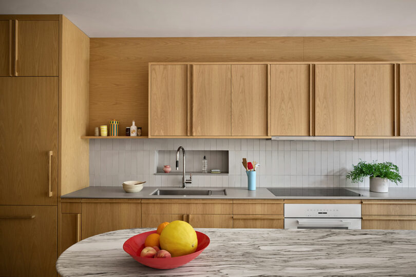 Modern kitchen with light wood cabinets, marble countertop with a red fruit bowl, stainless steel sink, white backsplash, and small potted plant near the stove.