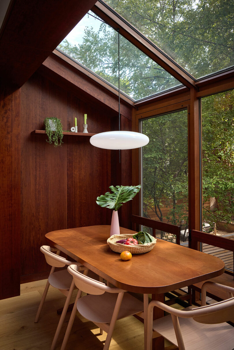 A wooden dining table with four chairs sits by large windows and a skylight. A white pendant light hangs above, with a vase, fruit bowl, and plant on the table. Trees are visible outside.