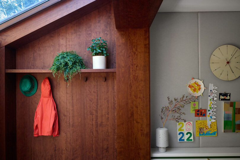 A wooden wall with two green hats, an orange jacket, and two potted plants; adjacent wall displays a clock, vase with branches, and colorful artwork.