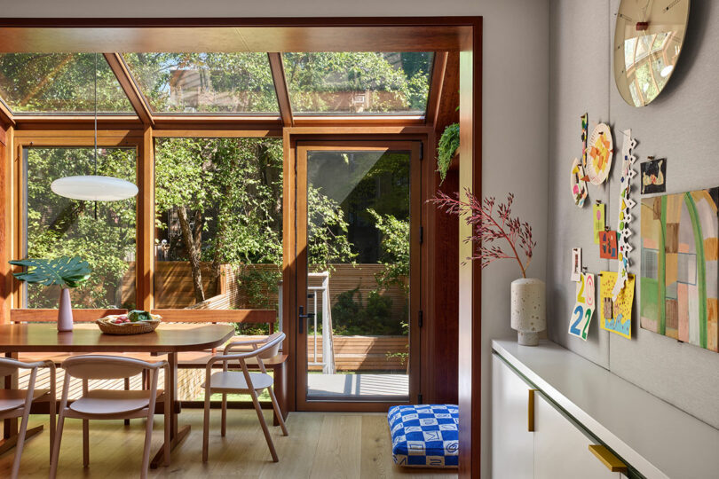 A modern dining area with wooden table and chairs is next to large glass doors and windows overlooking a green backyard. Children's artwork is displayed on a nearby wall.