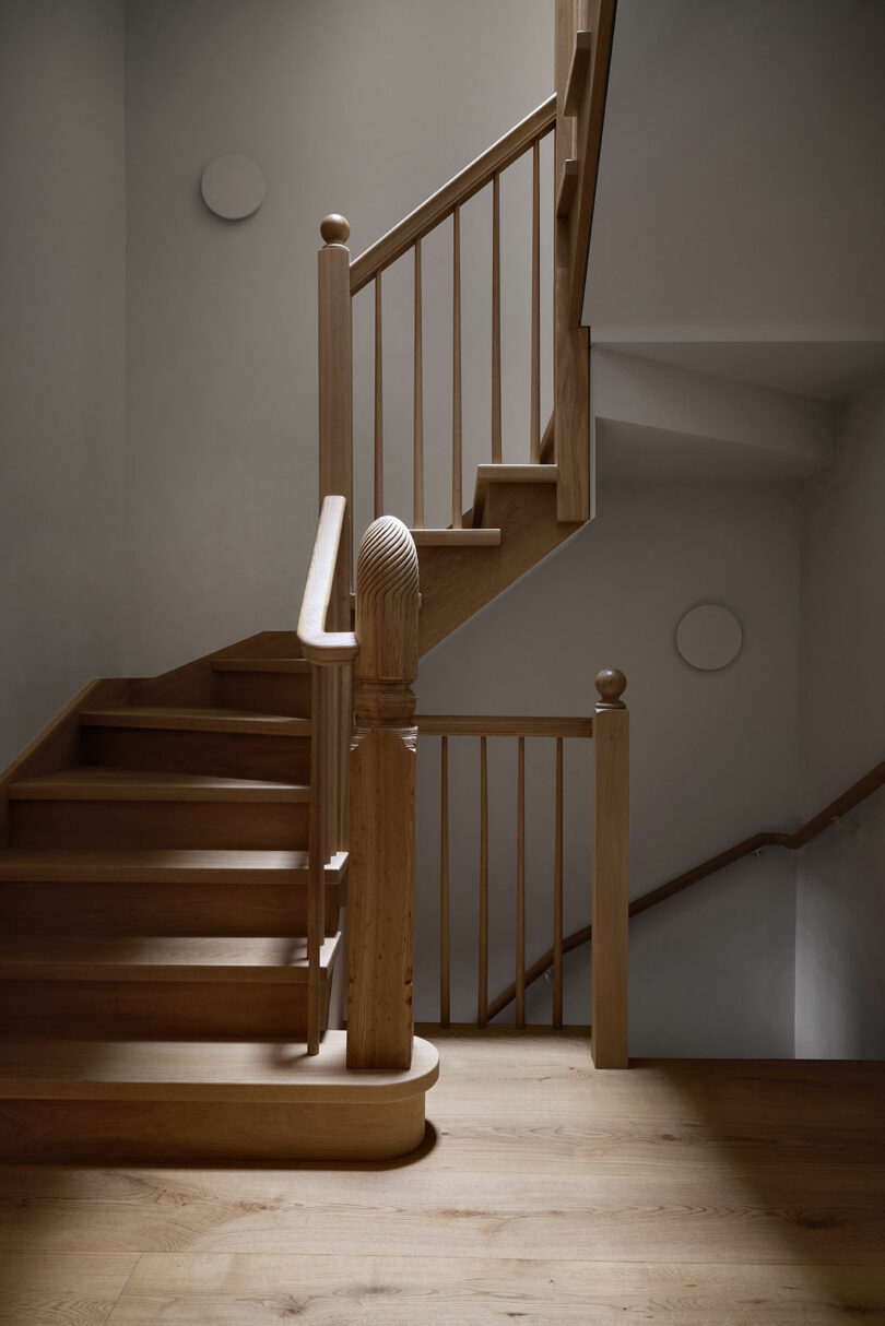Wooden staircase with simple railings and rounded posts, set against plain white walls in a well-lit interior space.
