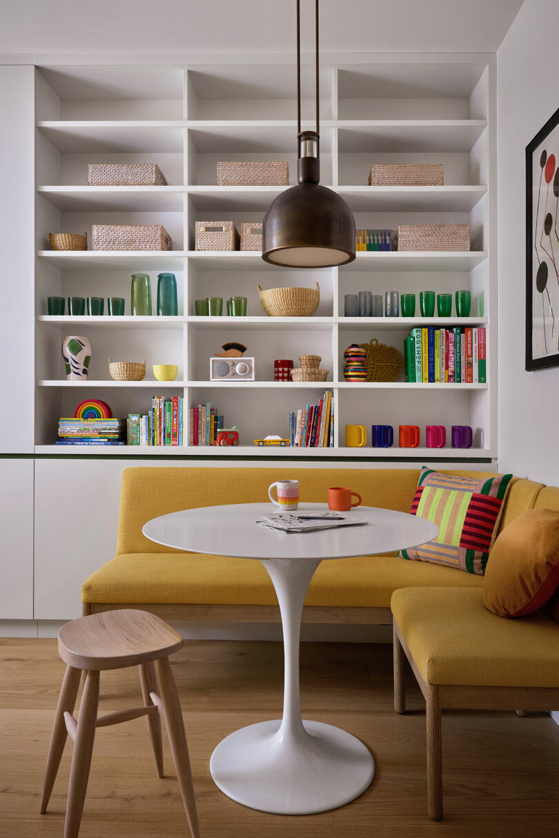 A modern dining nook with a yellow upholstered bench, white round table, wooden stool, colorful mugs, books, and decorative items on open shelves against a white wall.