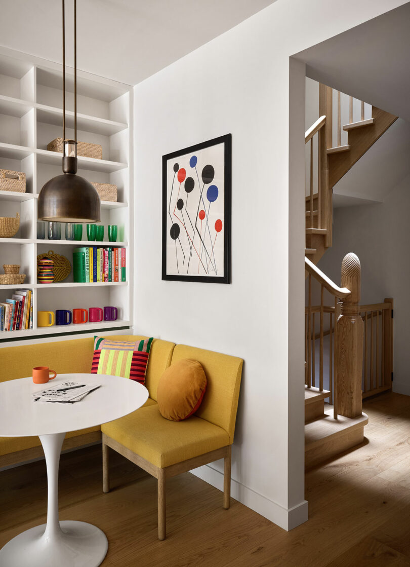 A modern dining nook with a yellow bench, round white table, colorful mugs, built-in shelves, abstract artwork, and a wooden staircase in the background.
